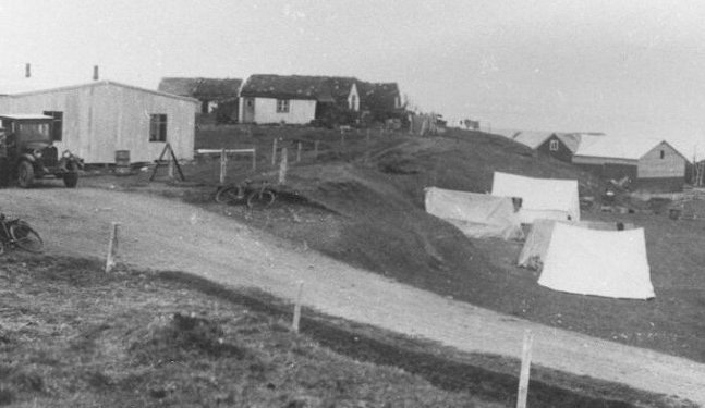 Úr Hvömmunum, norðan Brimnesár. Frá v.  Bráðabirgðahús sem reist var og rekið sem mötuneyti fyrir Dalvíkinga jarðskjálftasumarið. Á bakkanum er  f.v. Baldvinsbúð, Árhóll, Hansabúð. Fyrir neðan bakkann eru sjóhús bræðranna frá Háagerði Jóhanns og Friðleifs Jóhannssona. Tjaldbúðir í forgrunni.